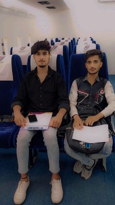 two young men sitting on blue seats in an airplane with white tables and chairs behind them