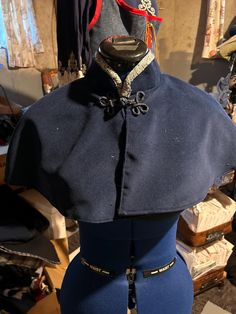 Overview: This beautifully crafted navy blue capelet is inspired by traditional military uniforms, featuring meticulous handmade details and a refined design. Material: Fabric: High-quality navy blue wool or wool-blend fabric, chosen for its durability, warmth, and elegant drape. Trim: Decorative metallic gold trim around the collar adds a touch of opulence and historical accuracy. Design Details: Front Closure: The capelet fastens at the front with decorative frog closures, providing both functionality and a classic military aesthetic. Collar: A high, structured collar adorned with gold trim enhances the regal and authoritative look of the piece. Cut and Shape: The capelet is designed to sit comfortably on the shoulders, flaring out slightly to allow for ease of movement while maintaining Royal Cape, Plague Doctor Costume, Costume Capes, Military Aesthetic, Shoulder Cape, Wool Cape, Military Uniforms, Elegant Drapes, Period Costumes