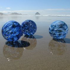 three blue glass balls sitting on top of a sandy beach next to the ocean with waves coming in