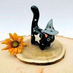 a black cat figurine sitting on top of a piece of wood next to a flower