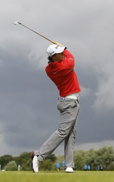 a man swinging a golf club on top of a lush green field with dark clouds in the background