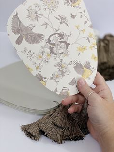 a hand holding a fan with flowers and birds printed on the side, next to a pair of scissors