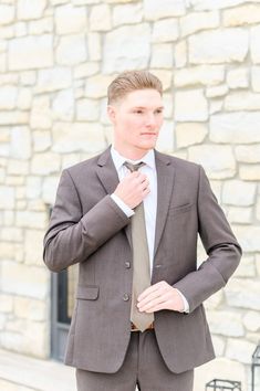 a young man adjusting his tie in front of a stone wall with a light colored suit