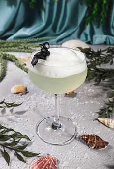 a glass filled with white liquid on top of a sandy beach next to sea shells