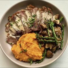 a white plate topped with meat and veggies next to mashed potatoes, asparagus and mushrooms