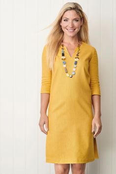 a woman standing in front of a white wall wearing a yellow dress and beaded necklace