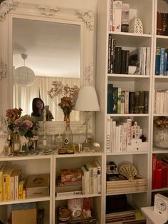 a white book shelf filled with books next to a large mirror and lamp on top of it