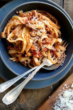 a blue bowl filled with pasta and sauce on top of a wooden table next to two silver spoons