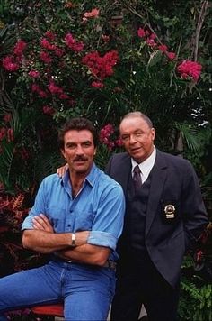 two men standing next to each other in front of trees and flowers with their arms crossed