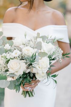 a bride holding a bouquet of white roses and greenery in her hands, wearing an off the shoulder wedding dress