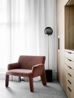 a pink chair sitting in front of a window next to a wooden cabinet and lamp