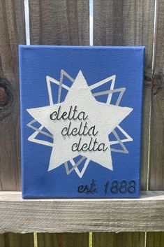 a blue and white canvas with the words delta delta delta on it sitting on a wooden fence