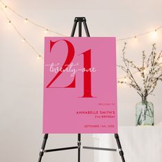 a pink 21st birthday party sign sitting on top of a easel next to a vase with flowers