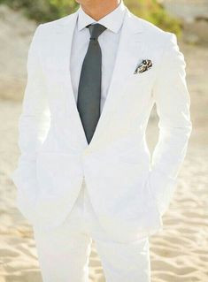 a man in a white suit and tie standing on the beach with his hands in his pockets