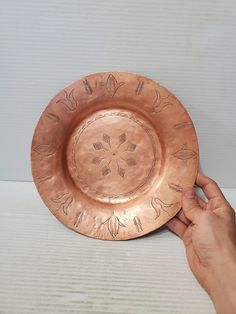 a hand is holding a copper plate with designs on it and the other hand is pointing at it