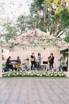 a group of people playing music under a tent with white flowers on the floor and greenery