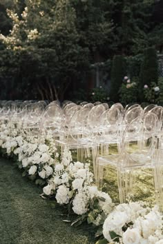 rows of clear chairs with white flowers on the grass in front of trees and bushes