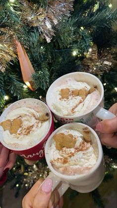 three people holding mugs filled with hot chocolate and marshmallows, next to a christmas tree