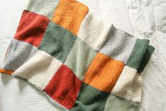 a blanket that is laying on top of a white bed sheet with an orange, green, and grey checkered design