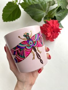 a woman's hand holding a pink coffee mug with a colorful dragonfly on it