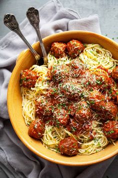 a bowl filled with spaghetti and meatballs on top of a gray cloth next to two spoons