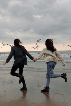 two people running on the beach with birds flying in the sky behind them and one person holding hands