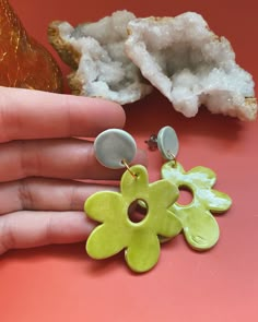 a person's hand next to two different colored flower shaped earrings on a pink surface