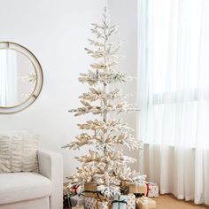 a white christmas tree with presents under it in front of a window and a round mirror