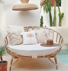 a wicker couch with pillows and a hat on it in front of a potted plant