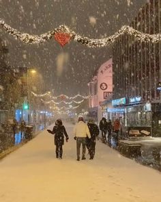 two people walking down a snowy street in the middle of winter with lights strung above them