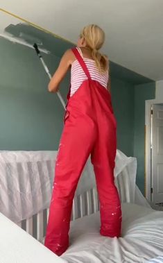 a woman in red overalls painting a wall with a paint roller on top of a bed