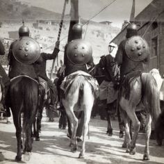 men in armor riding on horses down a dirt road with other people standing behind them