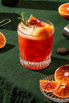 an orange cocktail garnished with rosemary sits on a green tablecloth next to sliced oranges