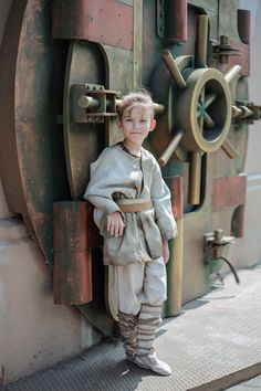 a young boy dressed in medieval clothing leaning against a door with a wheel on it