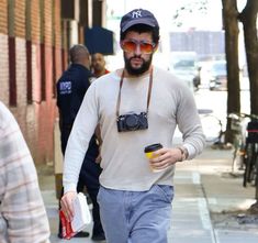 a man walking down the street with a camera on his shoulder and a drink in his hand