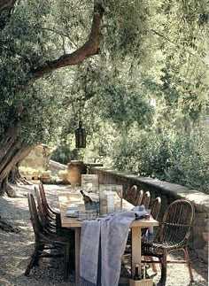 an outdoor table and chairs under trees on the side of a stone wall with water in it