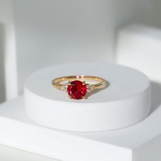 a gold ring with a red stone in the center on a white stand, sitting on a white surface