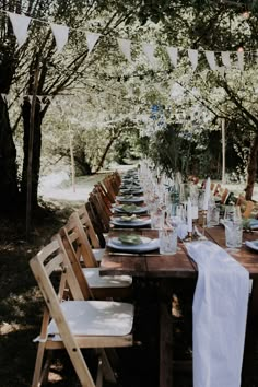 a long table set up with plates and glasses for an outdoor dinner in the woods