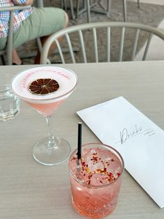 two cocktails sitting on top of a wooden table next to a paper and pen