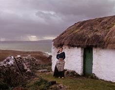 a woman standing in front of a thatched roof house next to the ocean on a cloudy day