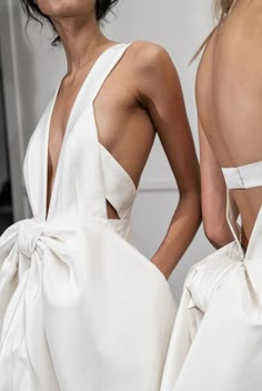 two models in white dresses with their backs turned to the side, one wearing a large bow