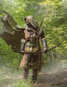 a man dressed in medieval clothing holding a bow and arrow while walking through the woods