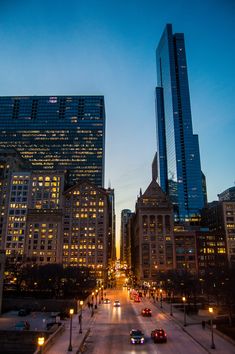 a city street at night with tall buildings and cars driving down the road in front of them