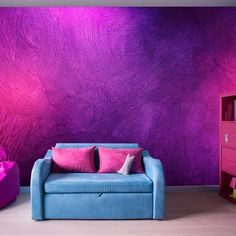 a blue couch sitting in front of a purple wall with pink pillows on top of it