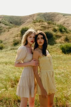 two young women standing next to each other in a field with grass and hills behind them