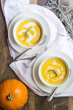 two white bowls filled with soup on top of a wooden table next to an orange pumpkin