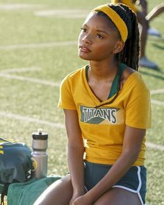 a young woman sitting on a bench in front of a sports field wearing a yellow shirt