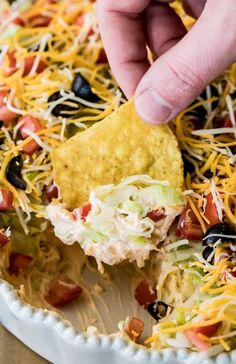 a person dipping a tortilla chip into a bowl full of shredded cheese and black olives