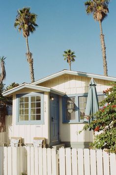 a small white house with palm trees in the back ground and a blue umbrella over the front door