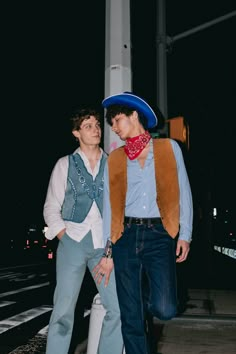 two young men standing next to each other on a city street at night, one wearing a cowboy hat